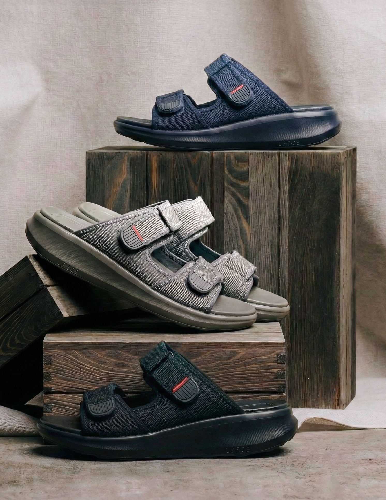 Three pairs of sandals in black, gray, and navy on wooden blocks against a beige fabric background.